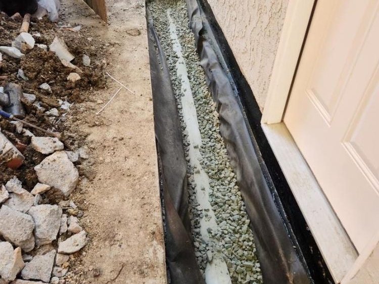 Drainage trench lined with membrane and filled with gravel beside a doorway